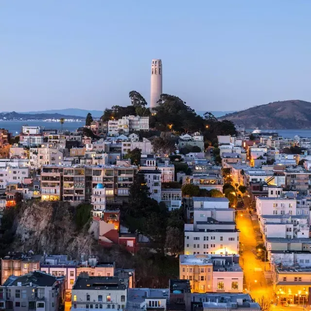 Coit Tower di San Francisco al tramonto, con le strade illuminate davanti e la baia di San Francisco alle spalle.