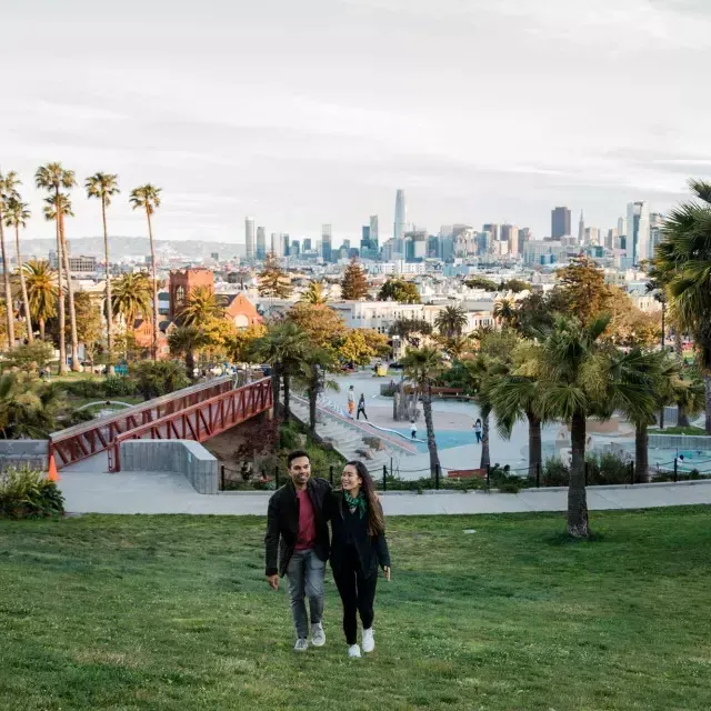 Una pareja camina hacia la cámara con Dolores Park y el horizonte de San Francisco detrás de ellos.